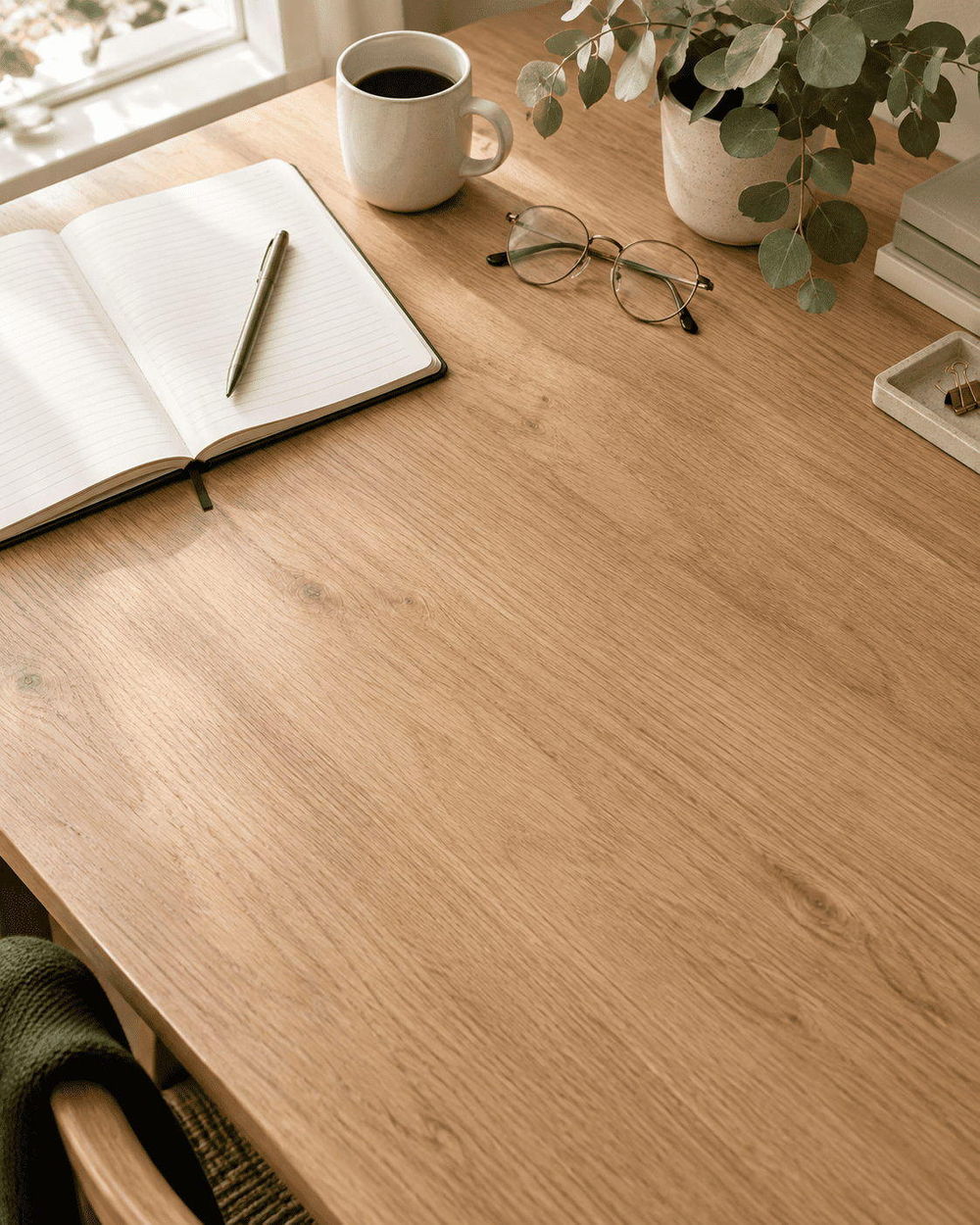 An oak studio desk seen from above: an open notebook and pen, a white mug, folded glasses and a potted eucalyptus, with the surface left clear below.